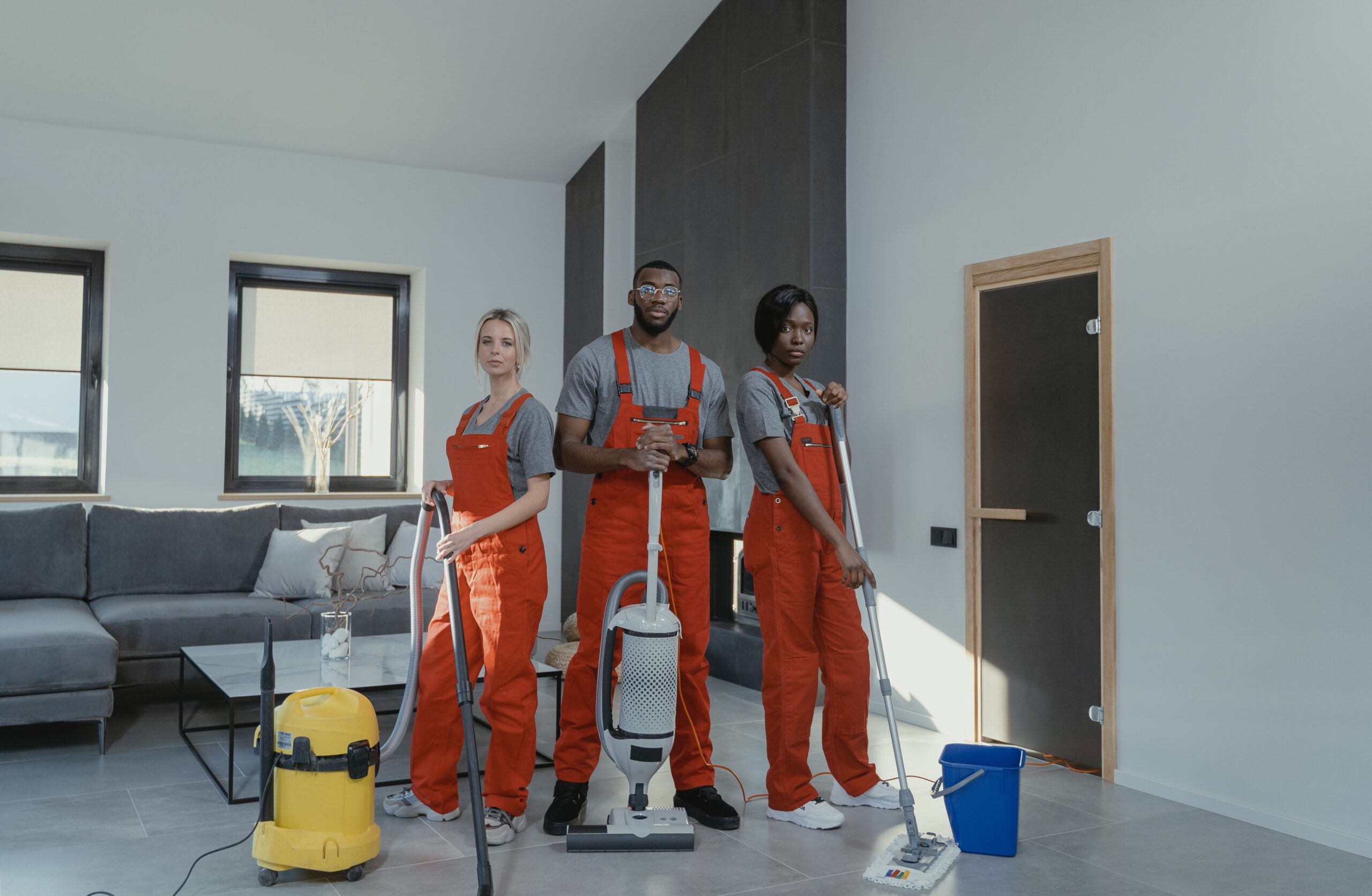 Inicio Diverse cleaning team in red uniforms ready to sanitize a modern living room with vacuum and mop.